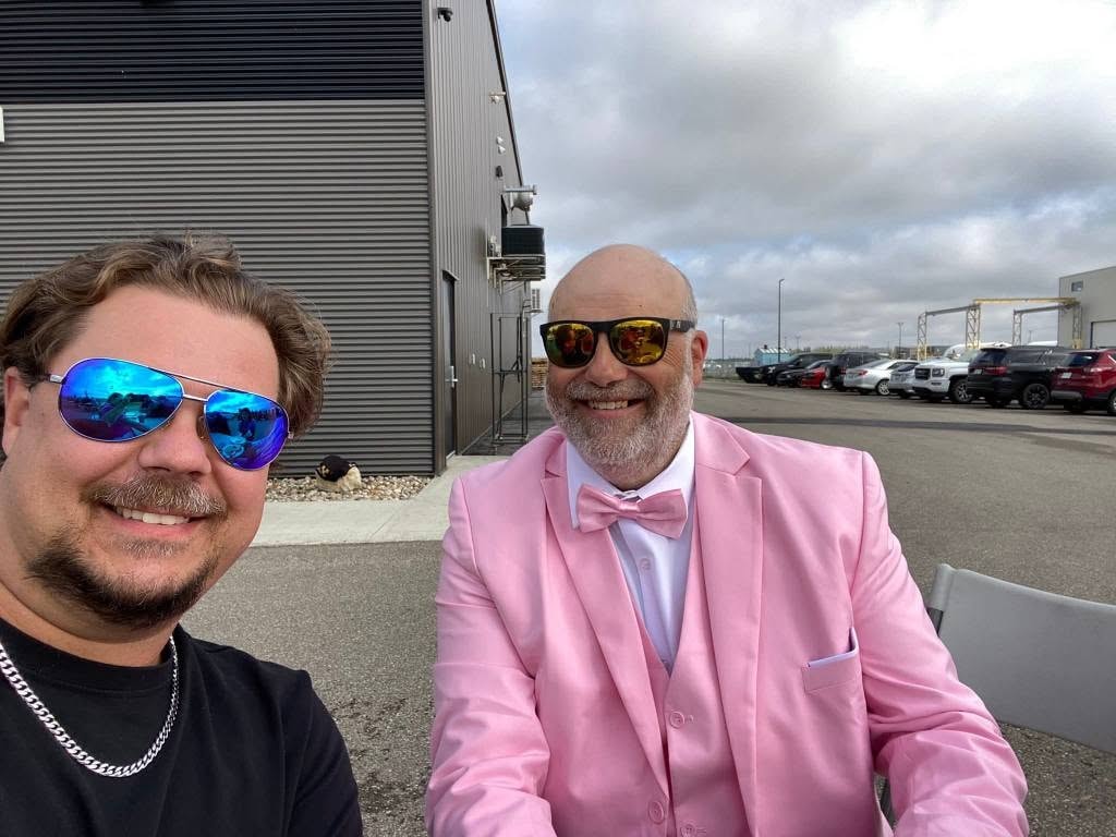 Two chapter members taking a selfie outside Prairie HD — one in a black t-shirt and blue aviators, one in a full pink three-piece suit and gold mirrored sunglasses at the 2025 Breast Cancer Awareness Ride