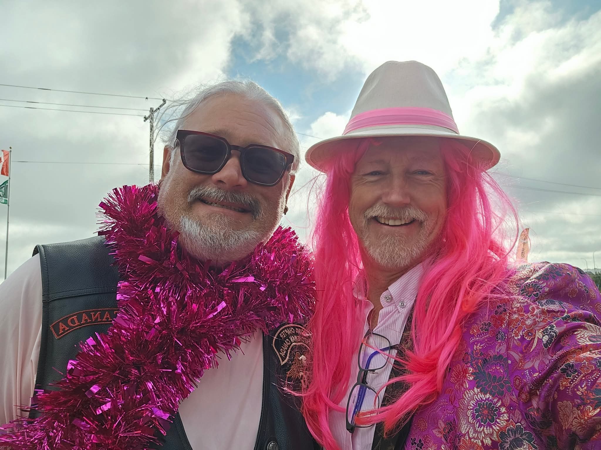 Two chapter members taking a selfie outdoors at the 2025 Breast Cancer Awareness Ride — one in a leather HOG vest with a hot-pink tinsel boa, one in a bright pink wig, white fedora and floral kimono, cloudy prairie sky behind them