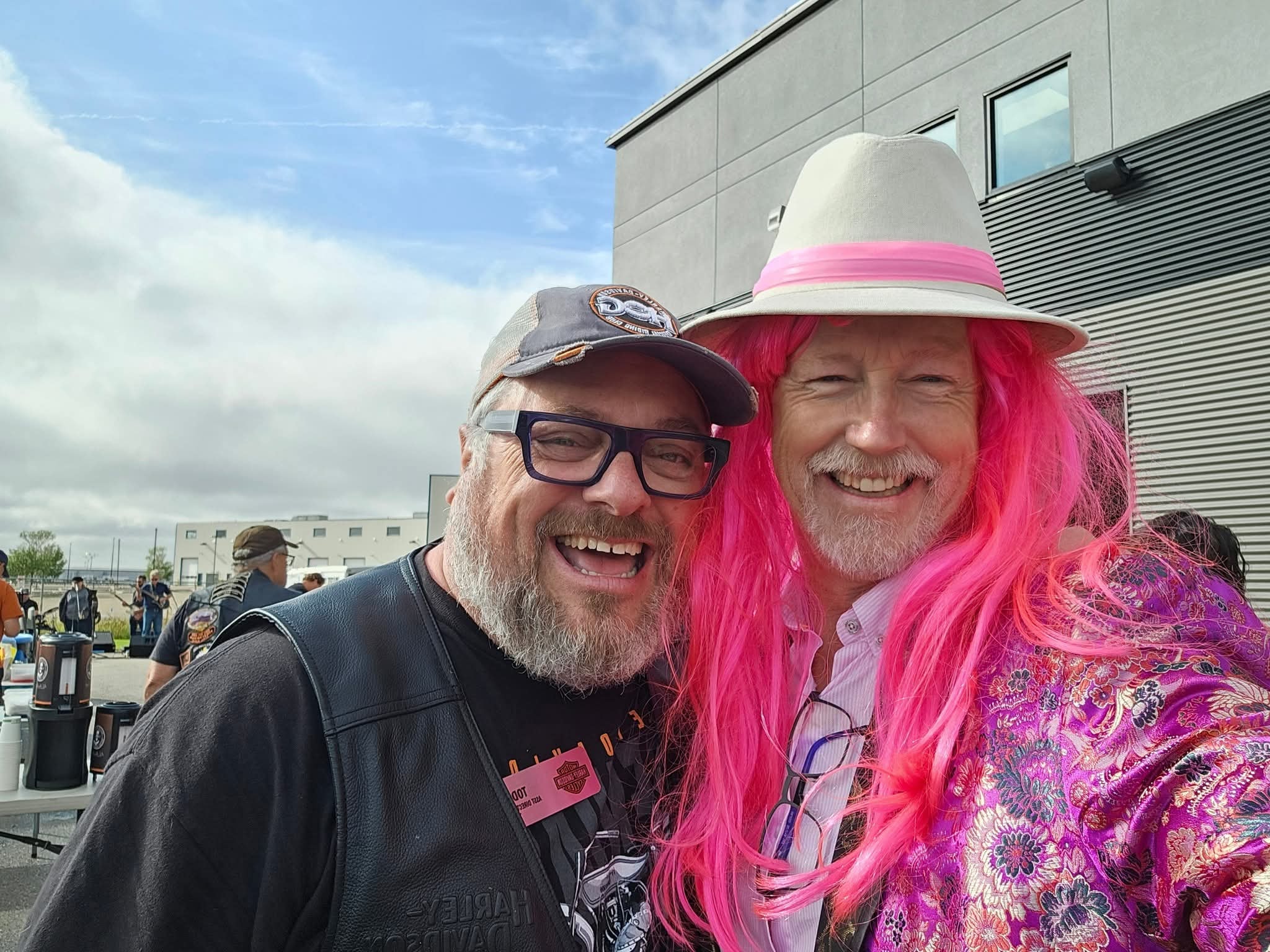 Two men laughing together outside Prairie Harley-Davidson at the 2025 Breast Cancer Awareness Ride — one in a Harley-Davidson cap and leather vest, one in a bright pink wig, white fedora and floral kimono