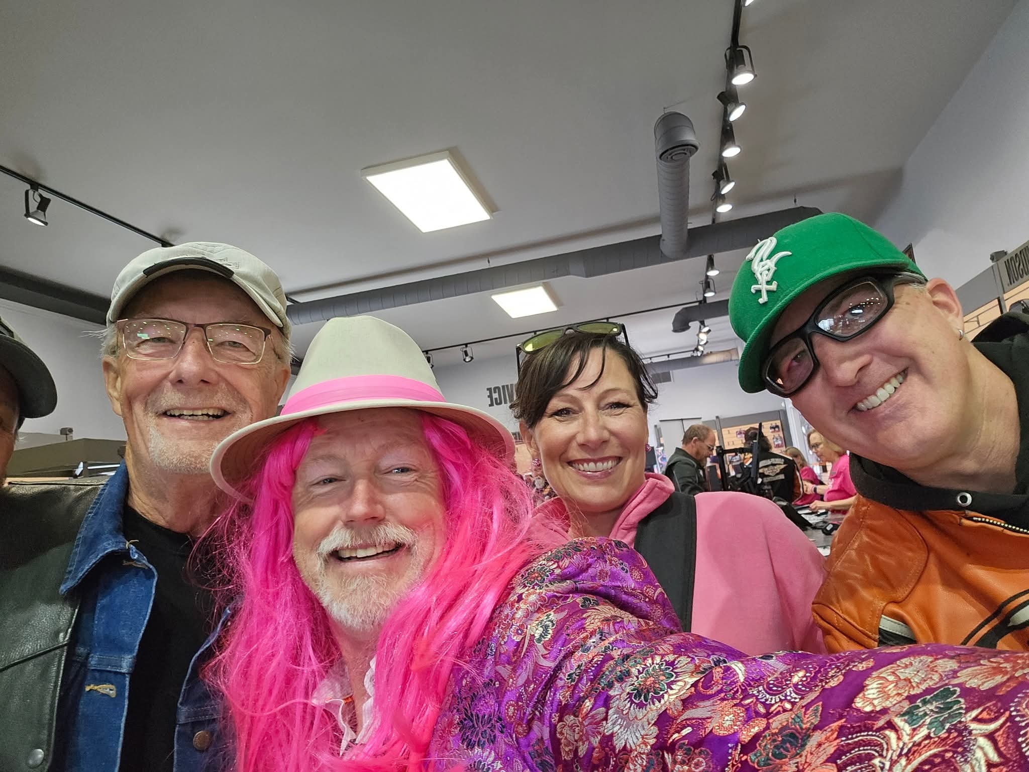 Four chapter members taking a group selfie inside the Prairie Harley-Davidson showroom at the 2025 Breast Cancer Awareness Ride — one in a pink wig and white fedora, one in a green cap and orange leather jacket, one in a denim jacket, and one woman in a pink jacket, with the busy event floor visible behind them