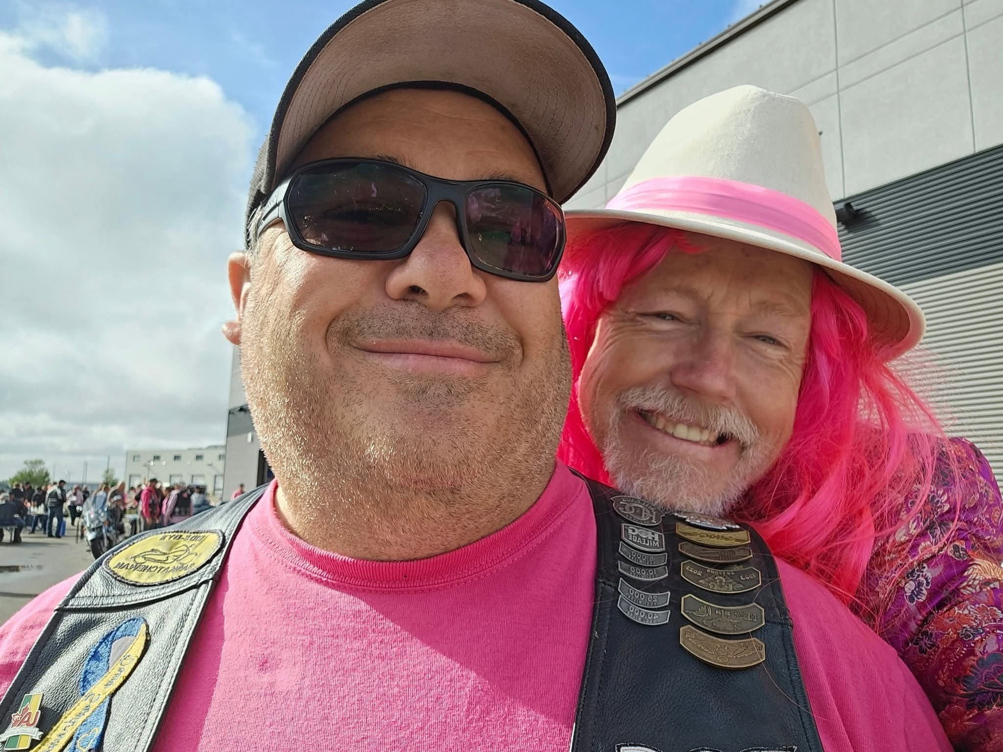 Two chapter members in pink — one in a pink shirt and HOG vest, one in a pink wig and white fedora — smiling outside Prairie Harley-Davidson at the 2025 Breast Cancer Awareness Ride
